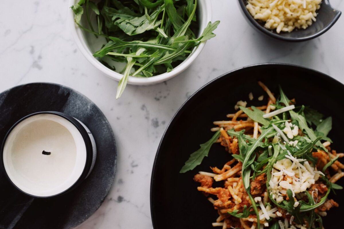 From above of plate with tasty spaghetti Bolognese sprinkled with fresh arugula and parmesan cheese placed on table with scented candle on black stand and bowls with greens and grated cheese